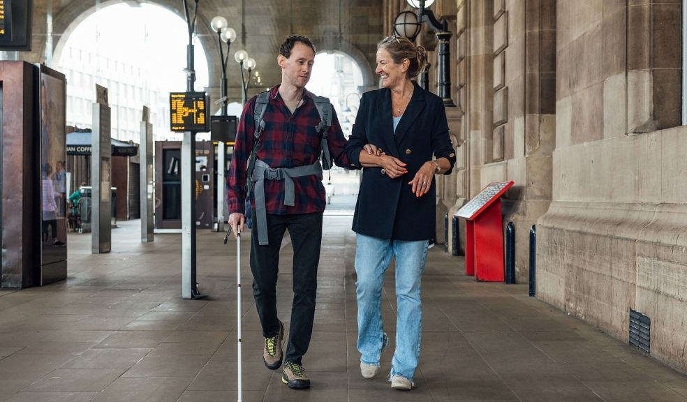 Visually impaired man with white walking aid walking through a station with a woman