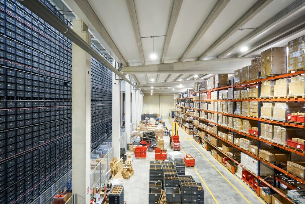 A large warehouse with tall storage racking, stacked boxes, pallets, and marked floor aisles