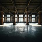 An empty industrial warehouse interior with brick columns, a concrete floor, and tall windows letting in light