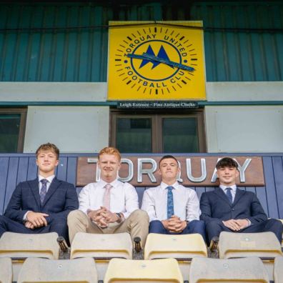 (Left to right) Toby Hard, Coby White, Fin Herbert and Jonah Shaw sit together in the stands at Torquay United Football Club, framed beneath the club’s crest. 