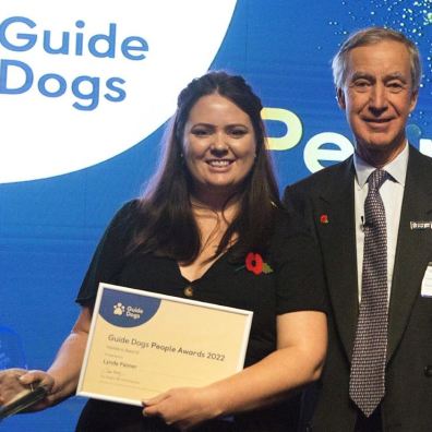 Lynda Palmer with her award standing with Jamie Hambro, Guide Dogs Chairman
