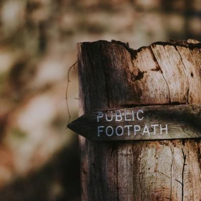 A photograph of a public footpath sign nailed to a tree stump, with woodland behind